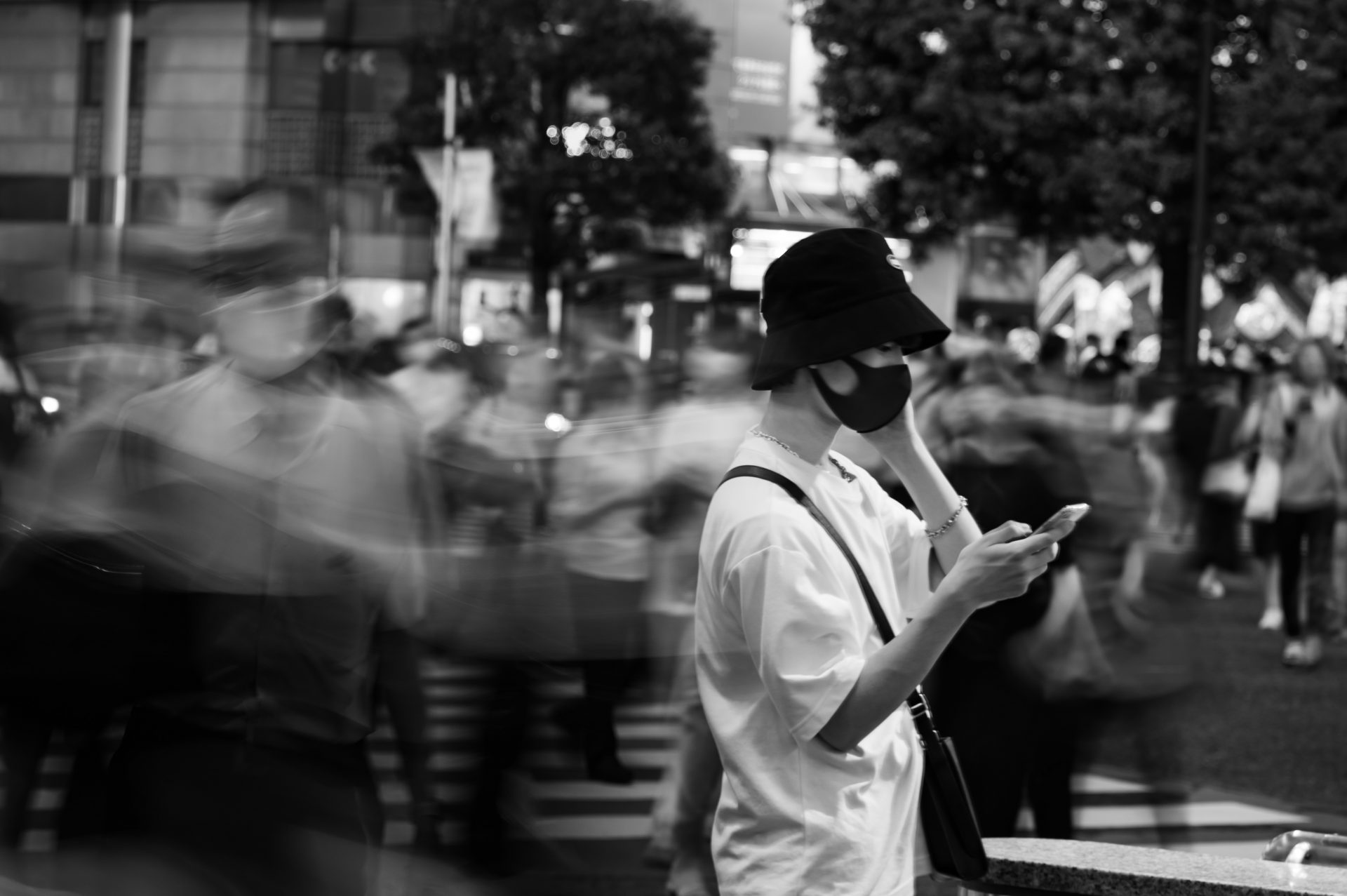 A boy texting on his phone while the pedestrians are passing by behind him. black and white street photography