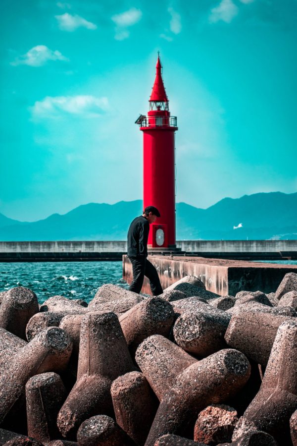 A man walking on the wavebreakers in the foreground in front of a red lighthouse